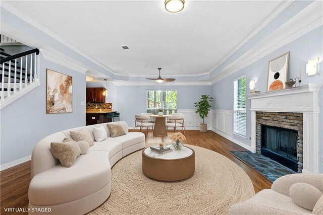 living room with wood finished floors, ceiling fan, a stone fireplace, crown molding, and a wainscoted wall