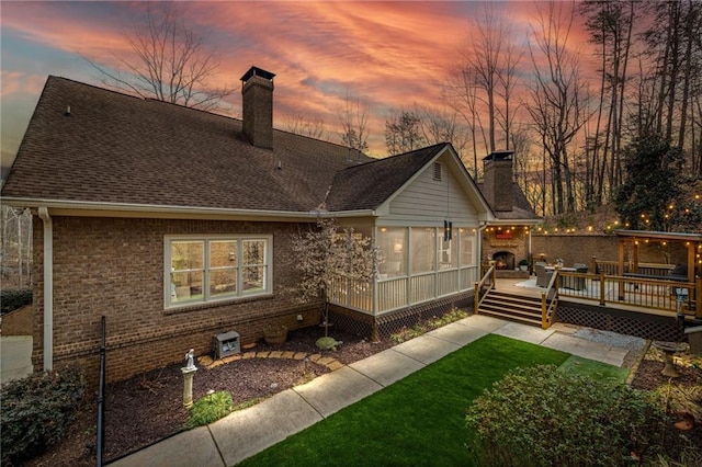 back of house at dusk with brick siding, a shingled roof, a lawn, a wooden deck, and a chimney