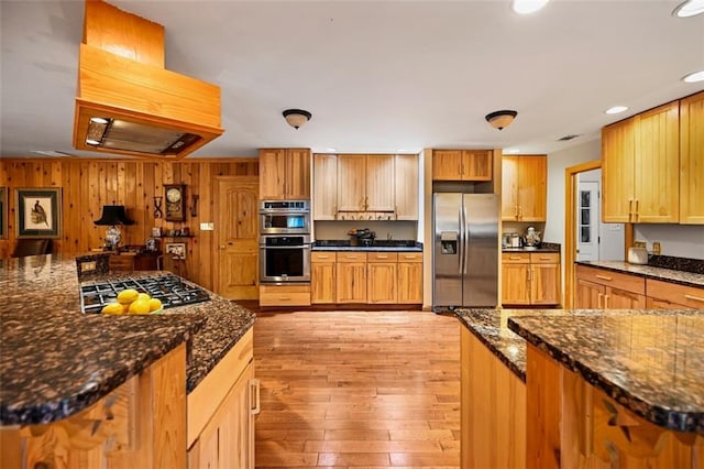 kitchen with wood walls, light hardwood / wood-style flooring, dark stone counters, island exhaust hood, and stainless steel appliances