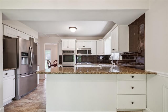 kitchen featuring appliances with stainless steel finishes, a sink, a peninsula, dark stone counters, and white cabinetry