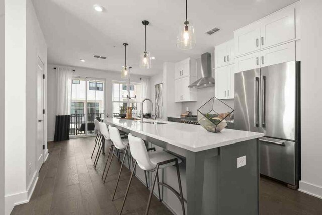 kitchen with high end refrigerator, dark hardwood / wood-style floors, an island with sink, wall chimney range hood, and white cabinets