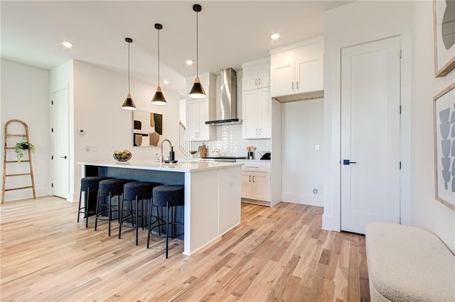 kitchen with decorative light fixtures, wall chimney range hood, white cabinetry, backsplash, and a center island with sink