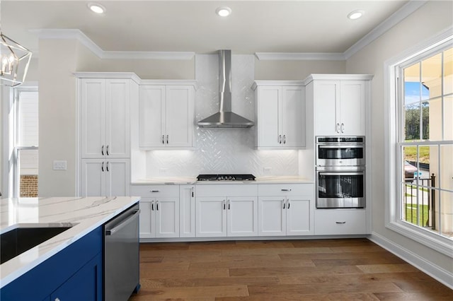 kitchen featuring blue cabinets, stainless steel appliances, ornamental molding, and wall chimney range hood