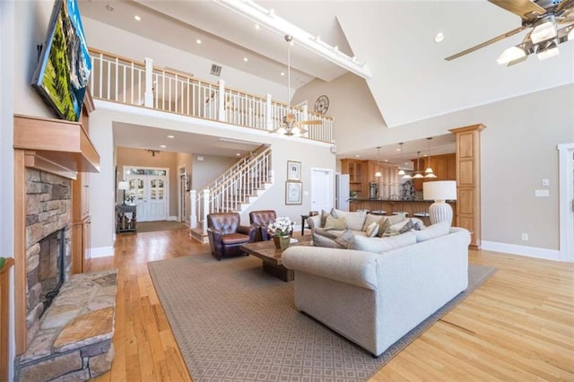 living area featuring light wood-style flooring, stairway, a high ceiling, a stone fireplace, and ceiling fan