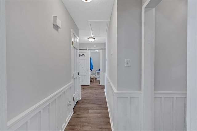 corridor with a textured ceiling, a barn door, a wainscoted wall, dark wood finished floors, and attic access