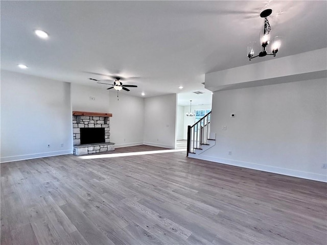 unfurnished living room with stairs, a stone fireplace, wood finished floors, baseboards, and ceiling fan with notable chandelier