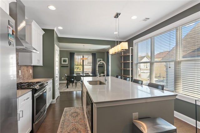 kitchen featuring an island with sink, appliances with stainless steel finishes, sink, wall chimney exhaust hood, and decorative light fixtures