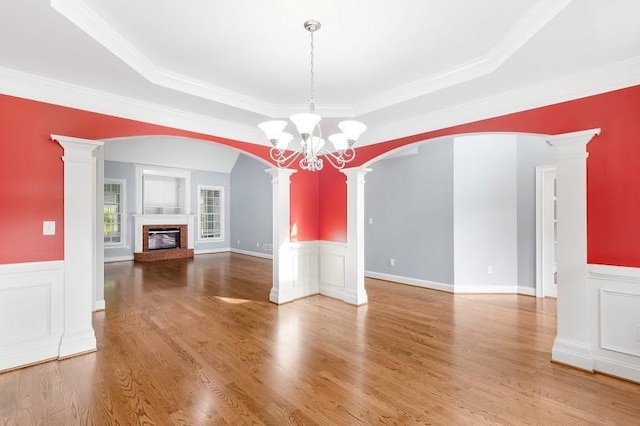 unfurnished living room featuring arched walkways, a fireplace, wood finished floors, a tray ceiling, and ornate columns