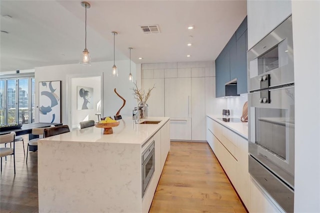 kitchen with light hardwood / wood-style floors, white cabinets, sink, an island with sink, and pendant lighting