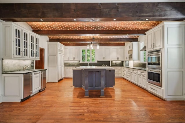 kitchen featuring stainless steel double oven, beamed ceiling, brick ceiling, light wood finished floors, and a kitchen island