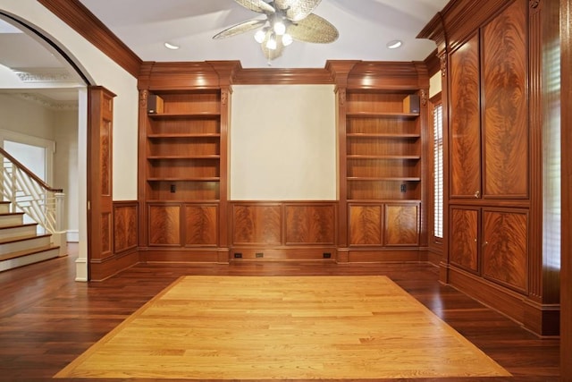unfurnished living room with ceiling fan, crown molding, arched walkways, built in features, and wainscoting