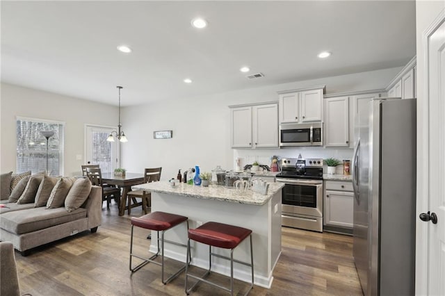 kitchen with decorative light fixtures, dark wood-type flooring, an island with sink, stainless steel appliances, and light stone counters