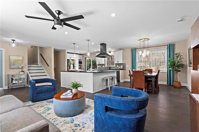 living area featuring stairway, baseboards, recessed lighting, dark wood-type flooring, and ceiling fan with notable chandelier