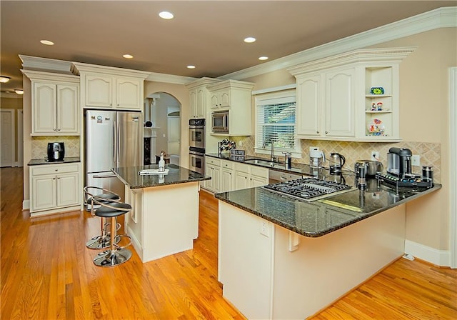 kitchen with arched walkways, dark stone countertops, a peninsula, stainless steel appliances, and light wood-style floors