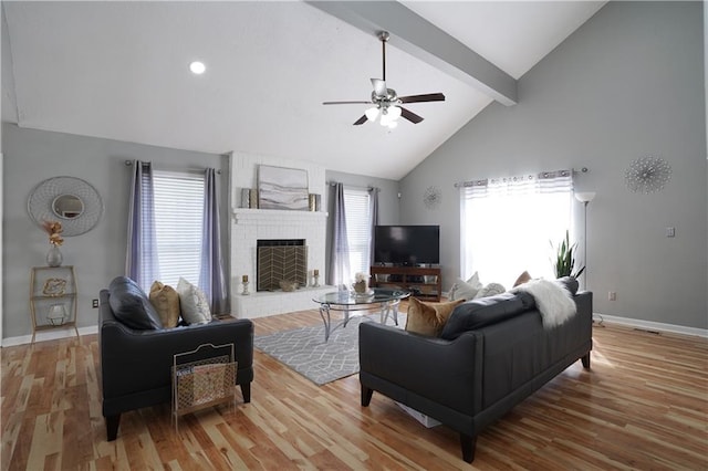 living room with high vaulted ceiling, a brick fireplace, light hardwood / wood-style flooring, ceiling fan, and beam ceiling