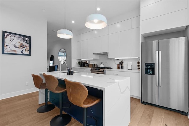 kitchen with pendant lighting, white cabinets, stainless steel appliances, light wood-type flooring, and a center island with sink