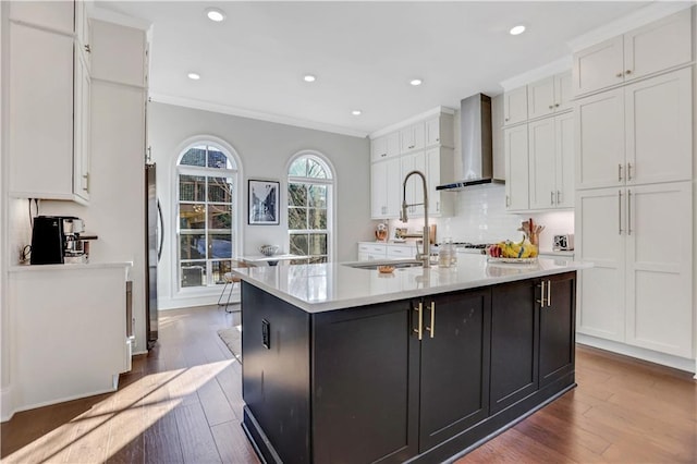 kitchen with white cabinetry, wall chimney range hood, crown molding, and an island with sink