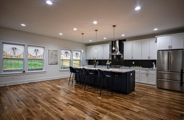 kitchen featuring pendant lighting, high end refrigerator, white cabinets, a kitchen island with sink, and wall chimney exhaust hood