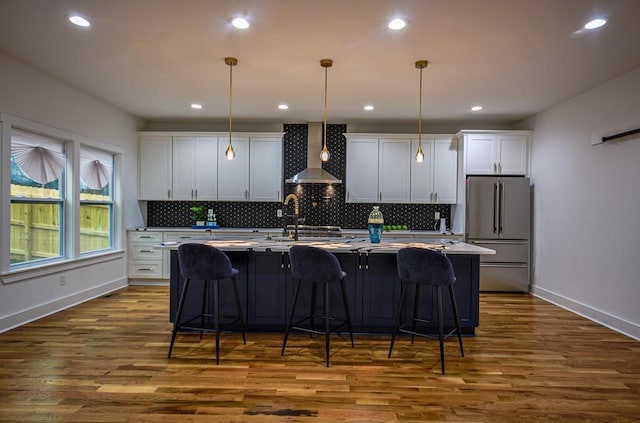 kitchen featuring wall chimney exhaust hood, decorative light fixtures, a center island with sink, stainless steel fridge, and white cabinets