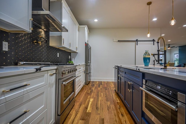 kitchen with appliances with stainless steel finishes, dark hardwood / wood-style floors, pendant lighting, white cabinets, and wall chimney exhaust hood