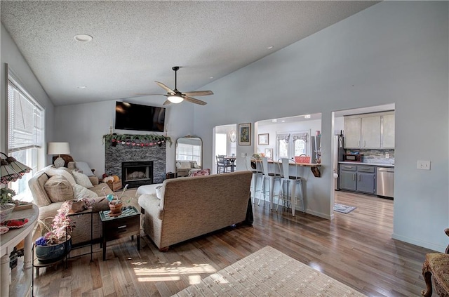 living area featuring a textured ceiling, a stone fireplace, light wood-style flooring, a ceiling fan, and baseboards