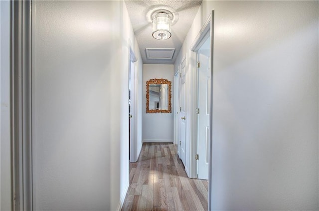 hallway with a textured ceiling, light wood finished floors, attic access, and baseboards