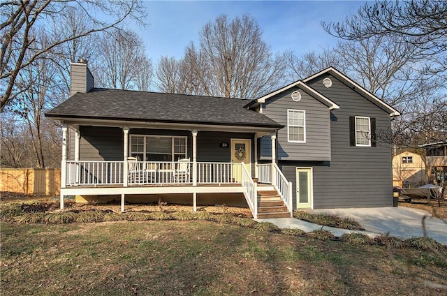 tri-level home with covered porch, a shingled roof, a chimney, and fence