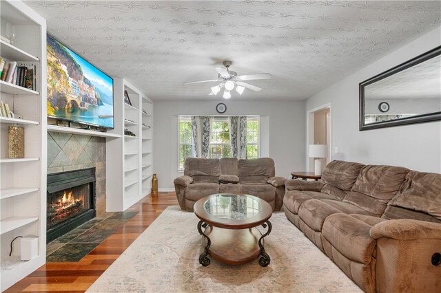 living room with dark wood-style flooring, a fireplace, a textured ceiling, built in shelves, and a ceiling fan