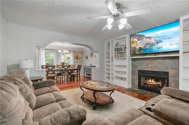 living room featuring arched walkways, built in shelves, wood finished floors, a tile fireplace, and ceiling fan with notable chandelier