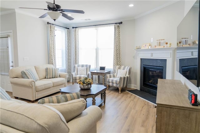 living area featuring visible vents, light wood-style flooring, a fireplace with flush hearth, ceiling fan, and crown molding