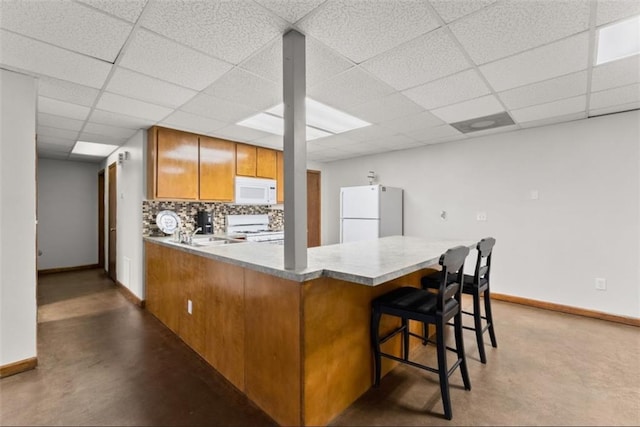 kitchen featuring white appliances, brown cabinetry, a peninsula, concrete flooring, and backsplash