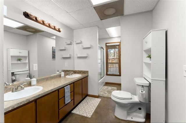 bathroom with double vanity, a drop ceiling, toilet, concrete flooring, and a sink