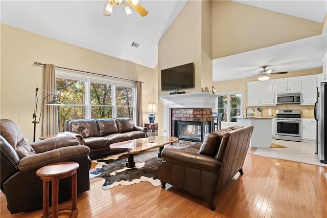 living room with a brick fireplace, high vaulted ceiling, light hardwood / wood-style flooring, and ceiling fan