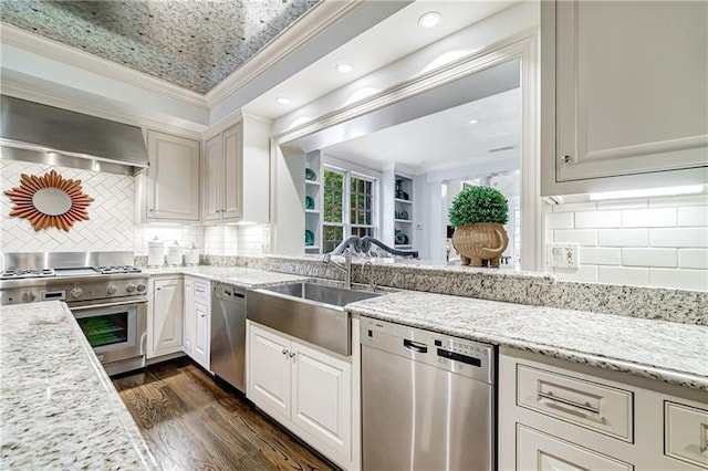 kitchen with appliances with stainless steel finishes, wall chimney range hood, a sink, ornamental molding, and dark wood finished floors