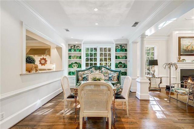 dining area with ornate columns, built in shelves, wood finished floors, crown molding, and a warm lit fireplace
