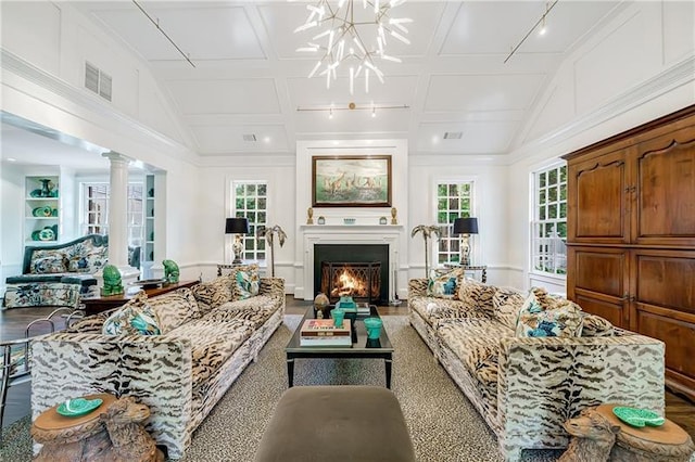 living room with a chandelier, a warm lit fireplace, vaulted ceiling, and ornate columns