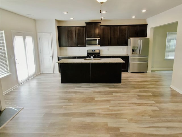 kitchen with appliances with stainless steel finishes, dark brown cabinets, a kitchen island with sink, and light stone countertops