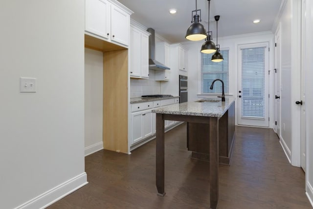 kitchen with pendant lighting, sink, wall chimney range hood, light stone counters, and a center island with sink