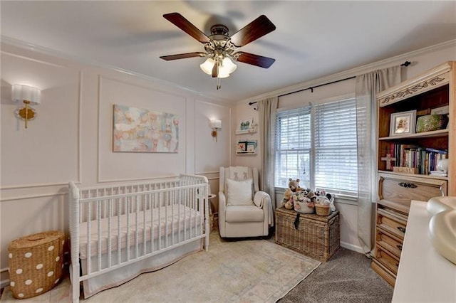 carpeted bedroom featuring a decorative wall, a crib, a ceiling fan, and ornamental molding