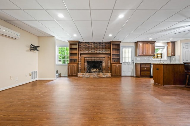 unfurnished living room with hardwood / wood-style flooring, a drop ceiling, an AC wall unit, and a brick fireplace