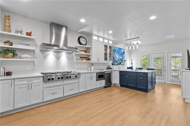 kitchen with a peninsula, light wood-type flooring, wall chimney range hood, open shelves, and appliances with stainless steel finishes