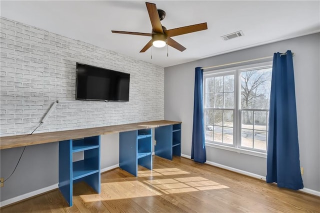 unfurnished office with ceiling fan, brick wall, wood finished floors, visible vents, and baseboards