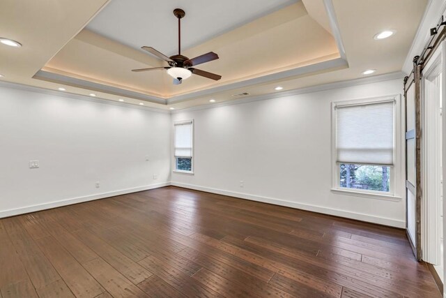 empty room featuring a tray ceiling, a ceiling fan, a barn door, dark wood-style floors, and baseboards