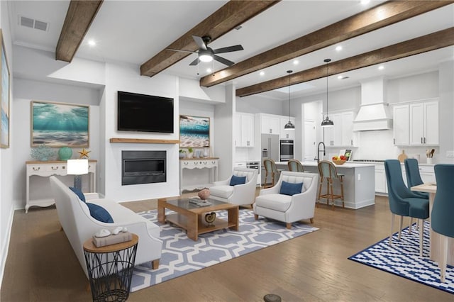 living area featuring visible vents, a glass covered fireplace, ceiling fan, wood finished floors, and beam ceiling