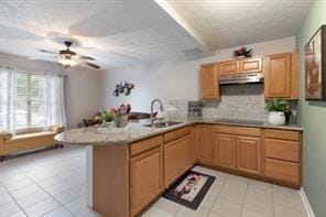 kitchen with a sink, a peninsula, under cabinet range hood, light tile patterned floors, and tasteful backsplash