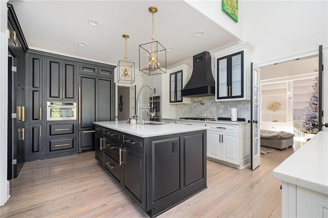 kitchen with custom exhaust hood, pendant lighting, a center island with sink, light hardwood / wood-style flooring, and white cabinets