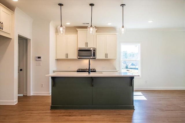 kitchen with wood finished floors, light countertops, stainless steel microwave, decorative light fixtures, and an island with sink