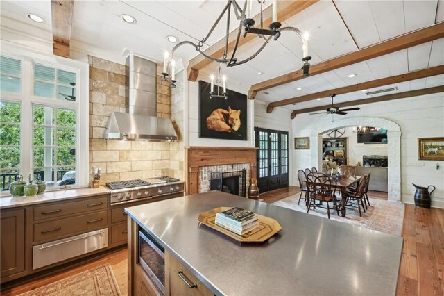 kitchen with light hardwood / wood-style floors, stainless steel counters, stainless steel microwave, a fireplace, and wall chimney exhaust hood