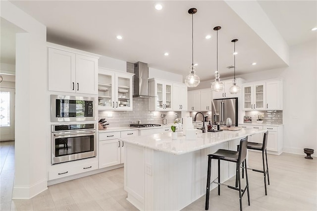 kitchen featuring wall chimney range hood, backsplash, stainless steel appliances, an island with sink, and decorative light fixtures