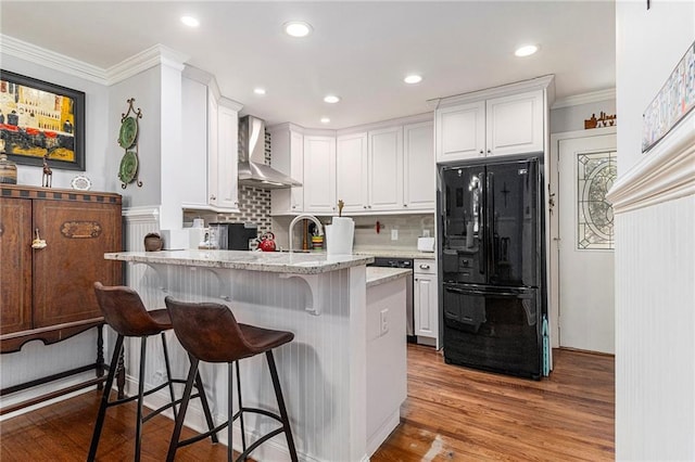 kitchen with light stone counters, a peninsula, white cabinetry, freestanding refrigerator, and wall chimney exhaust hood